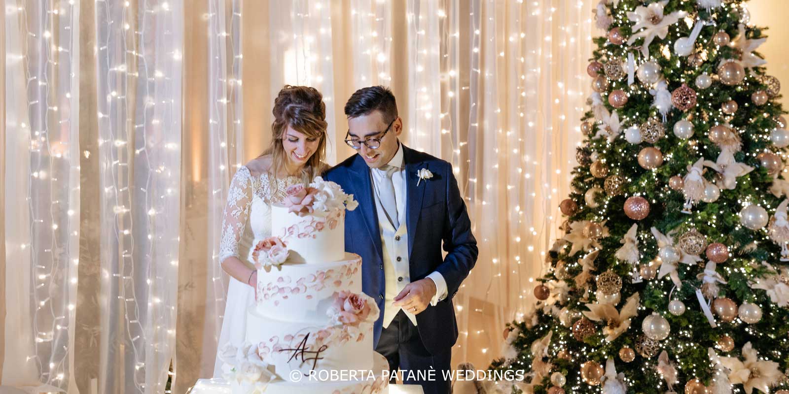 Matrimonio-a-tema-natalizio_Roberta-Patane-Weddings_2429 Sposi che tagliano la torta in un matrimonio a tema natalizio con l'albero di Natale e tenda di luci di sfondo. Un matrimonio firmato Roberta Patanè Weddings.